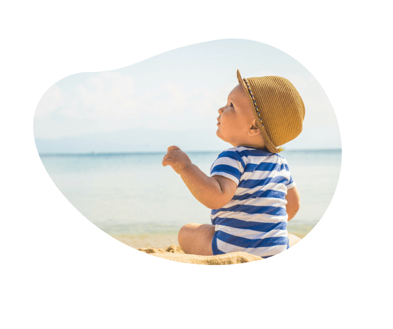 Kleinkind sitzt am Meer im blau weiss gestreiften Body und Strohhut.